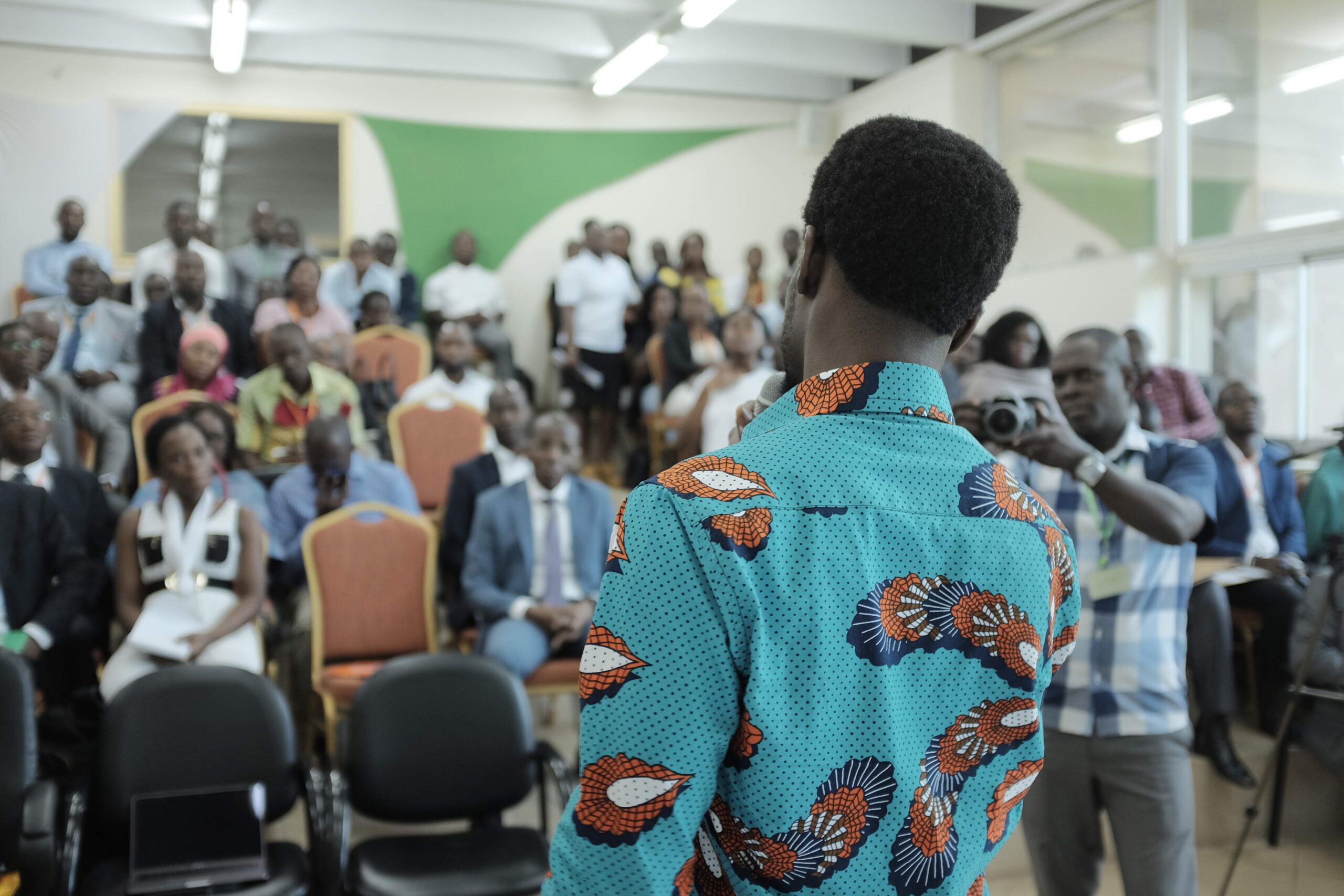 Man in a colourful shirt seen from behind, talking to a busty room