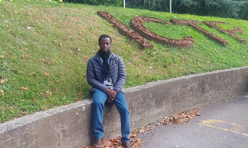 Romaric sitting on a grass verge near giant ICTP letters